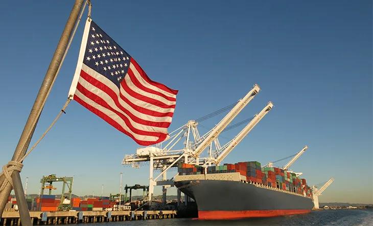 Le drapeau américain flotte à l’avant-plan dans un port américain, où un cargo chargé de conteneurs est amarré sous des grues géantes par une journée au ciel complètement dégagé.